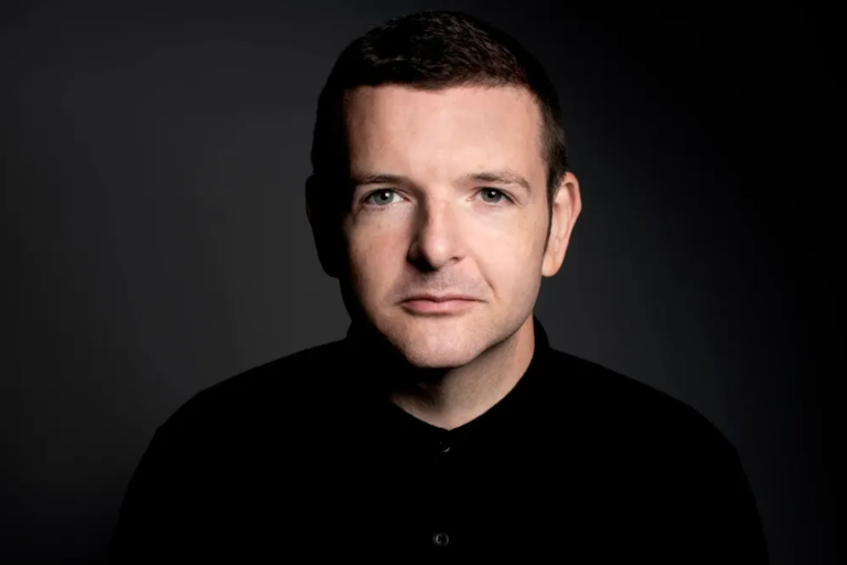 Close-up promo portrait of Scottish stand-up comedian Kevin Bridges wearing a black shirt against a dark studio background looking forward.